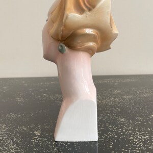 May include: A ceramic bust of a woman with a light brown hairdo and a green earring. The bust is painted in shades of pink and white.