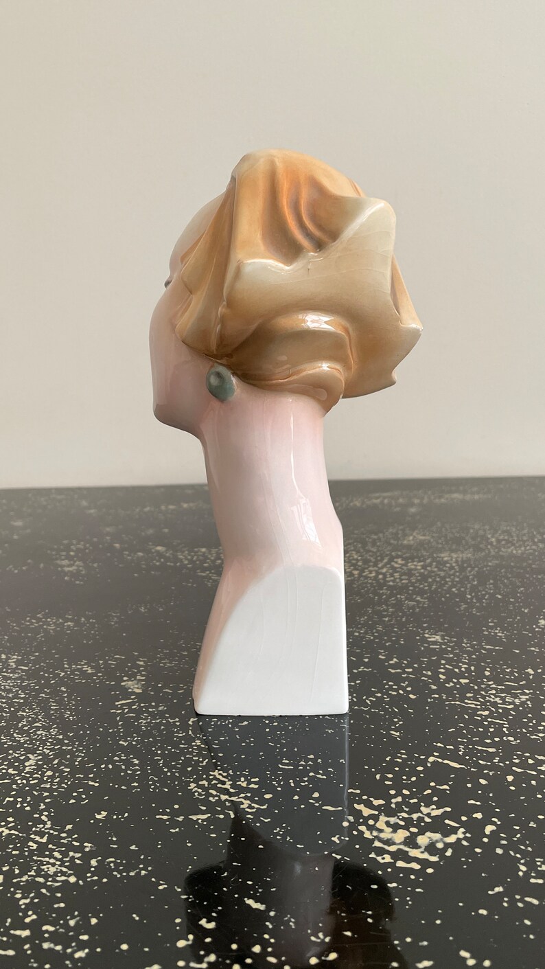 May include: A ceramic bust of a woman with a light brown hairdo and a green earring. The bust is painted in shades of pink and white.