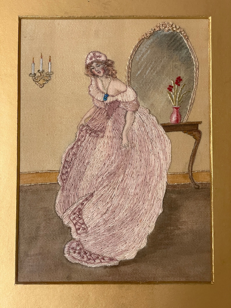 Puede incluir: Una mujer con un vestido rosa y un gorro blanco se encuentra de pie frente a un espejo. Est&aacute; mirando hacia abajo a su vestido. Hay un jarr&oacute;n con flores rojas sobre una mesa junto al espejo.