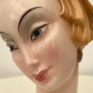 May include: A ceramic bust of a woman with a light brown hairstyle, painted blue eyes, and a pink blush on her cheeks. The bust is painted in a realistic style.