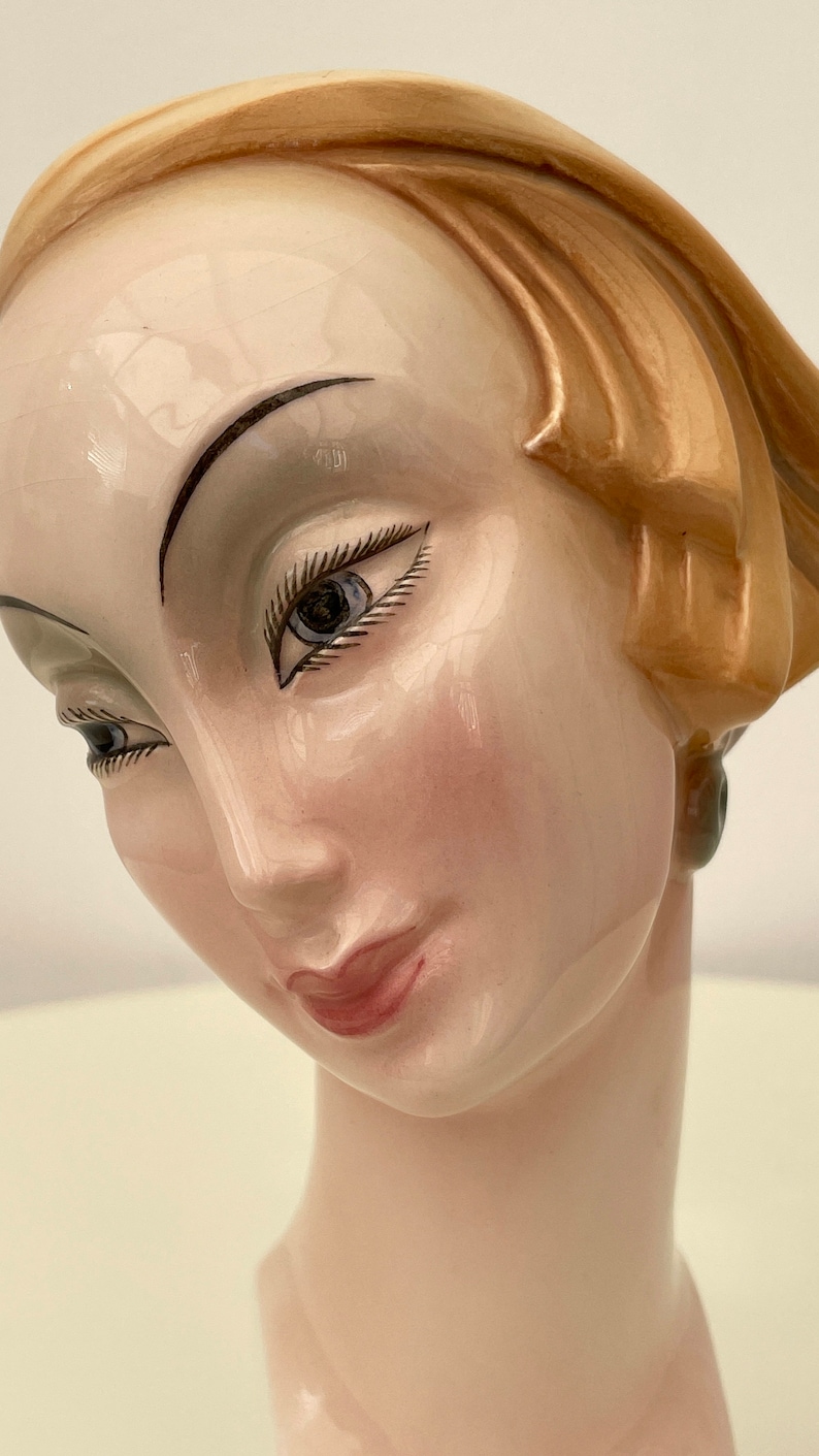 May include: A ceramic bust of a woman with a light brown hairstyle, painted blue eyes, and a pink blush on her cheeks. The bust is painted in a realistic style.