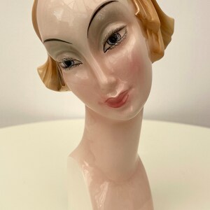 May include: A ceramic bust of a woman with a light brown hairstyle. The bust is painted with realistic features, including brown eyes, a pink mouth, and a light pink blush on the cheeks. The bust is on a white pedestal.