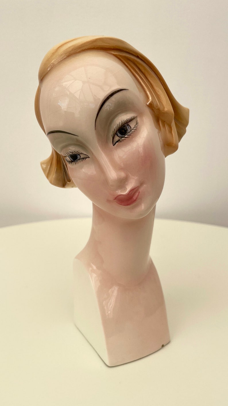 May include: A ceramic bust of a woman with a light brown hairstyle. The bust is painted with realistic features, including brown eyes, a pink mouth, and a light pink blush on the cheeks. The bust is on a white pedestal.