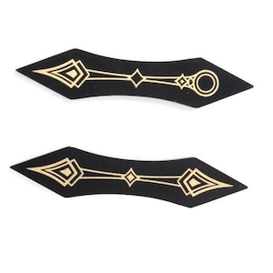 May include: Two black, curved appliques with gold geometric designs. Each applique has a pointed end and a series of diamond and circle shapes. The appliques are symmetrical and appear to be made of a soft material.