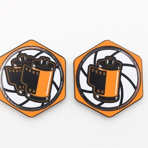 May include: Two orange hexagonal enamel pins with a white and black film roll design. The film roll is inside a circle with a white and black camera shutter design.