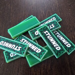 May include: A pile of green game tiles with the word "STUNNED" printed on them in white letters. The tiles have a black and white pattern.