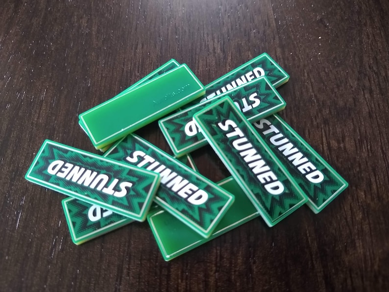 May include: A pile of green game tiles with the word "STUNNED" printed on them in white letters. The tiles have a black and white pattern.