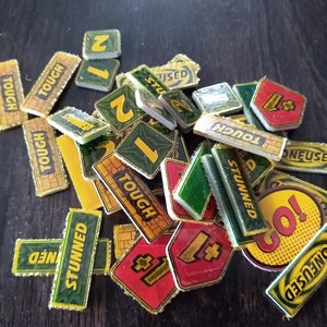 May include: A collection of game pieces for a board game. The pieces are made of cardboard and have various colours, including green, yellow, red, and black. The pieces feature words like "Tough", "Stunned", "Confused", and "Go!" as well as numbers.