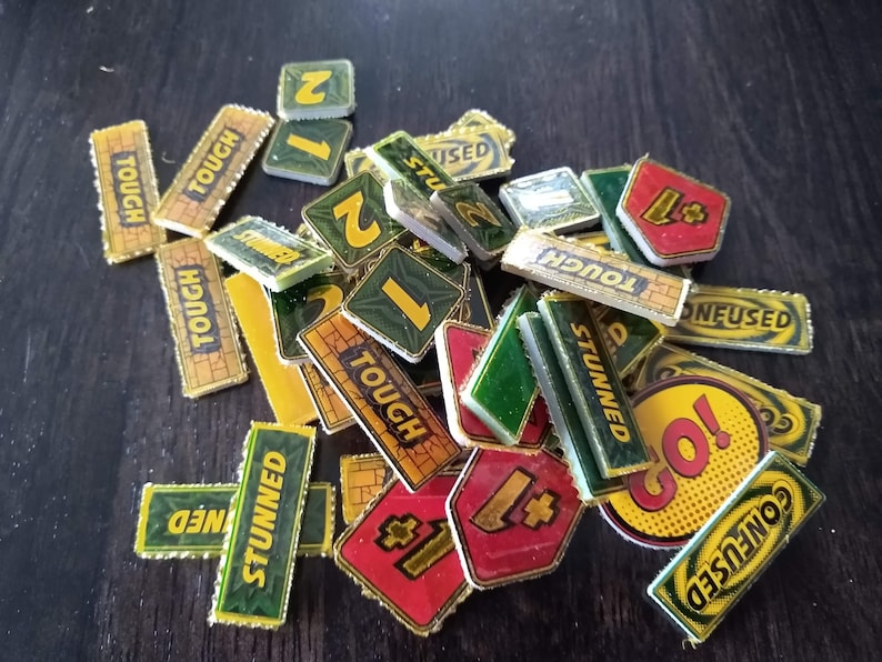 May include: A collection of game pieces for a board game. The pieces are made of cardboard and have various colours, including green, yellow, red, and black. The pieces feature words like "Tough", "Stunned", "Confused", and "Go!" as well as numbers.