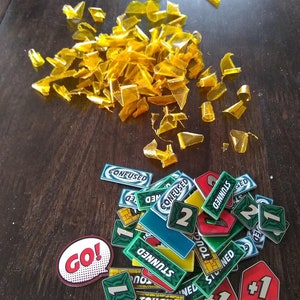 May include: A collection of colourful game pieces for a board game. The pieces are made of plastic and have various shapes and designs. Some of the pieces have numbers on them, while others have words like "Go!" and "Confused".