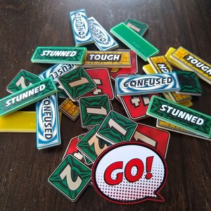 May include: A collection of green, blue and yellow game tiles with words such as "Confused", "Stunned", and "Tough" printed on them. There are also several green tiles with the number "1" printed on them. A red and white "GO!" speech bubble is in the centre of the image.