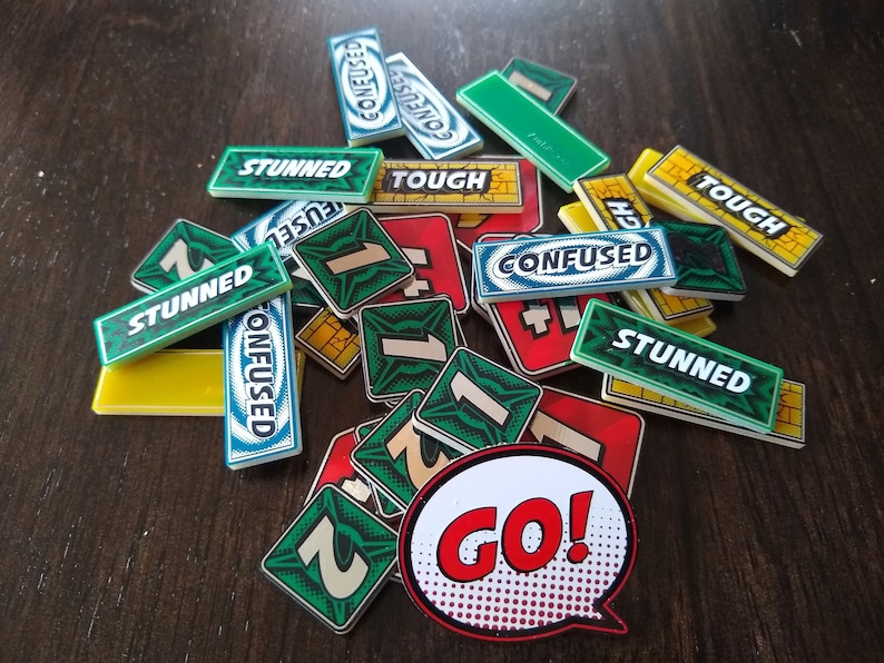 May include: A collection of green, blue and yellow game tiles with words such as "Confused", "Stunned", and "Tough" printed on them. There are also several green tiles with the number "1" printed on them. A red and white "GO!" speech bubble is in the centre of the image.