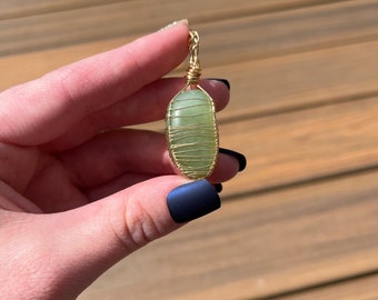 May include: A green stone pendant wrapped in gold wire. The pendant is oval shaped and hangs from a gold chain.