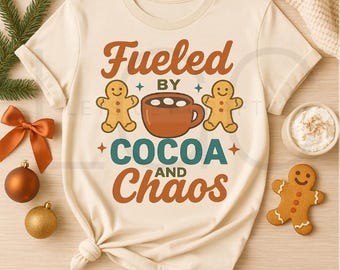 May include: Cream-colored t-shirt with the phrase "Fueled by Cocoa and Chaos" in brown, teal, and orange. The design includes gingerbread men and a cocoa mug. A festive and cozy graphic tee.