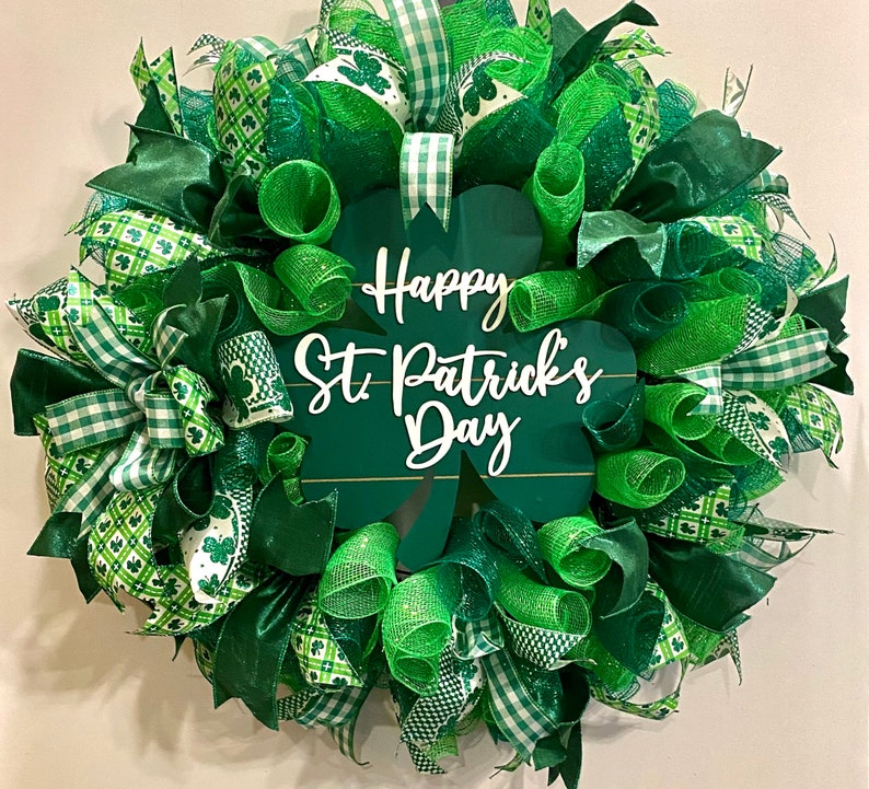 May include: A green and white St. Patrick's Day wreath with a shamrock-shaped sign that reads 'Happy St. Patrick's Day'. The wreath is made of green mesh, green and white checkered ribbon, and green and white shamrock patterned ribbon.