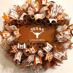 May include: A brown and white wreath with orange and white ribbons. The wreath features a brown metal sign with the words "Texas Longhorns" in white lettering.