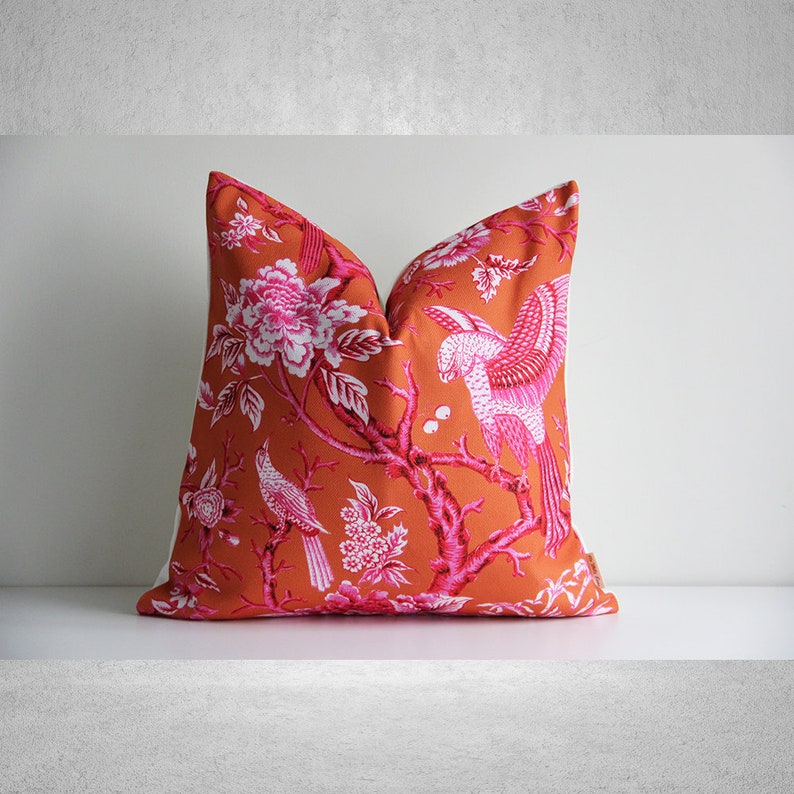 May include: Orange pillow with a pink floral pattern featuring birds. The pillow has a white border.