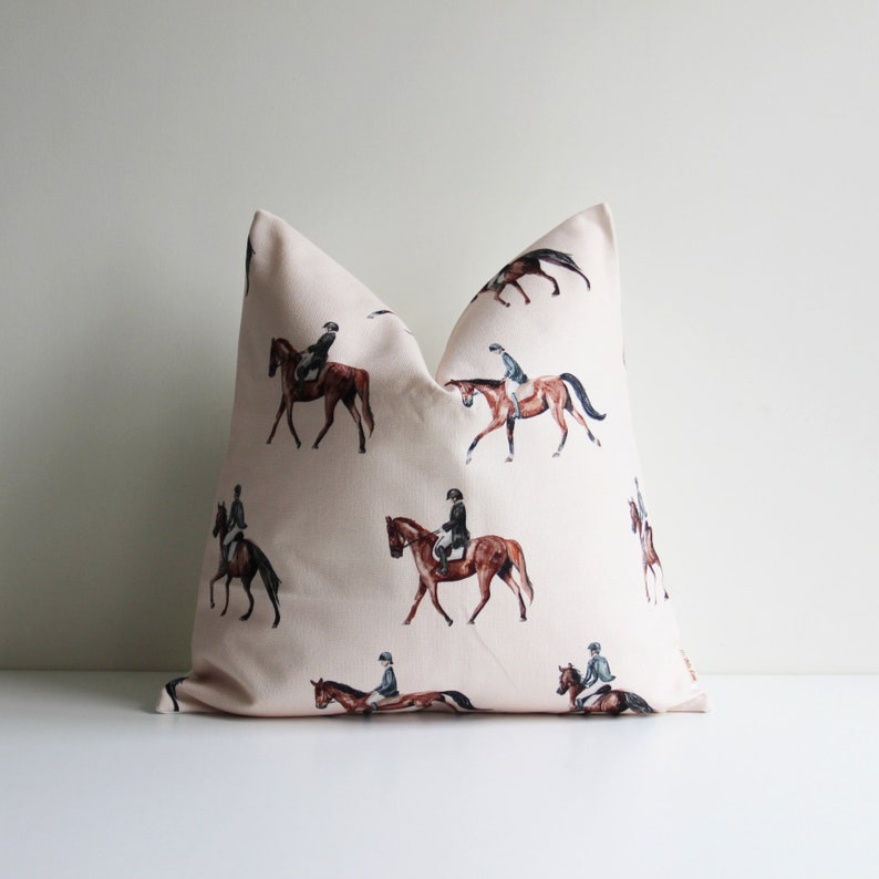 May include: A cream coloured pillow with a repeating pattern of brown horses and riders in blue and white clothing.