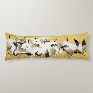 以下が含まれることがあります： 黄色い背景に白い鶴が飛んでいるデザインの長い長方形の枕。鶴は伝統的な日本のスタイルで描かれています。