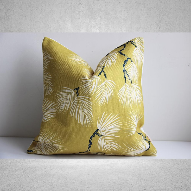 May include: Mustard yellow throw pillow with a white and navy blue palm leaf pattern. The pillow is square and has a soft, textured appearance. The design features stylized palm fronds.