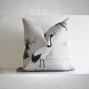 May include: A beige throw pillow with a black and white illustration of a crane standing in a field of grass. The crane has a red beak and black and white feathers. The background is a light beige with a subtle texture.