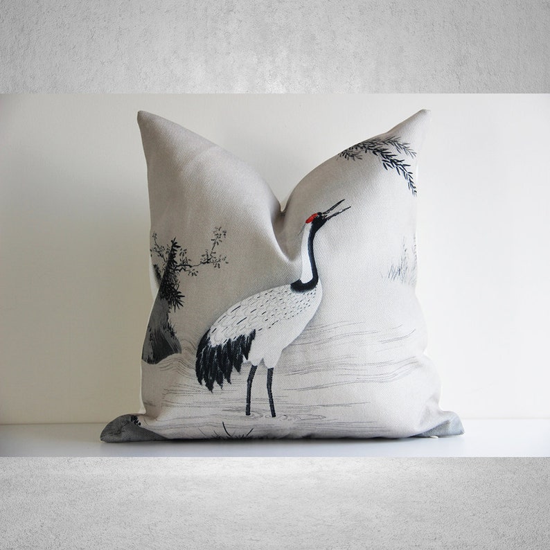 May include: A beige throw pillow with a black and white illustration of a crane standing in a field of grass. The crane has a red beak and black and white feathers. The background is a light beige with a subtle texture.