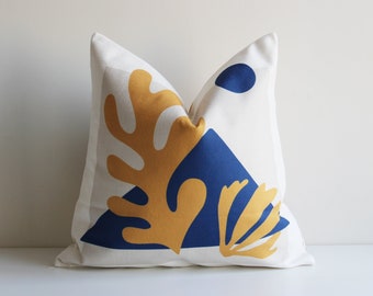 Puede incluir: Una almohada blanca con un diseño geométrico que presenta un triángulo azul, una hoja amarilla y un círculo azul. La almohada está hecha de una tela suave y natural.