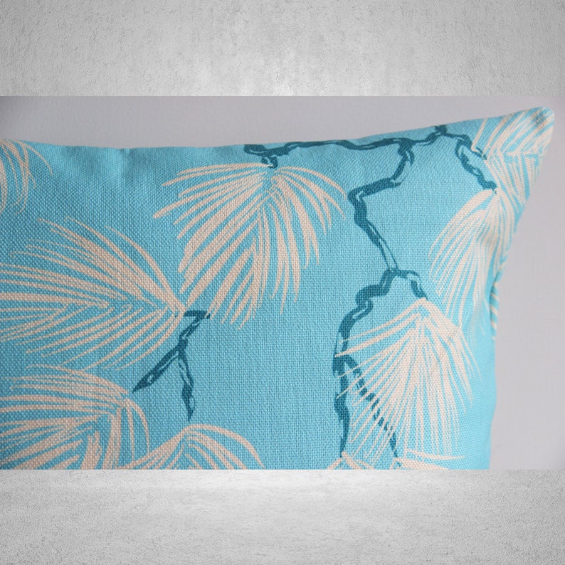 May include: A rectangular turquoise pillow with a repeating pattern of off-white palm leaves and dark blue stems. The pillow has a textured appearance, suggesting a linen or similar fabric. The design evokes a tropical or coastal theme.