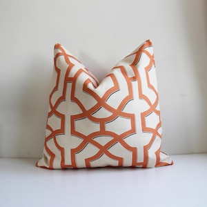 Könnte beinhalten: Ein quadratisches Zierkissen mit geometrischem Muster. Das Kissen hat einen cremefarbenen Hintergrund mit einem sich wiederholenden Muster aus orangefarbenen, ineinandergreifenden Formen, die schwarz umrandet sind. Es ist aus Stoff gefertigt und dient Dekorationszwecken.