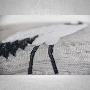 May include: A close-up of a gray and white throw pillow with a black embroidered design of a bird with long legs.
