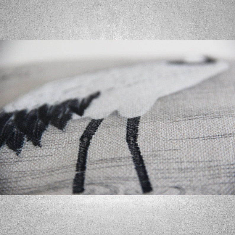 May include: A close-up of a gray and white throw pillow with a black embroidered design of a bird with long legs.