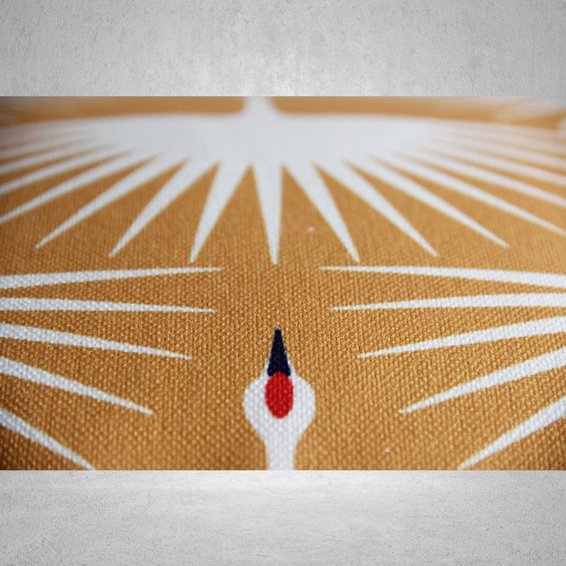 May include: Close-up of a brown fabric cushion with a white crane design. The crane's beak is red and black, and its wings are spread out in a starburst pattern.