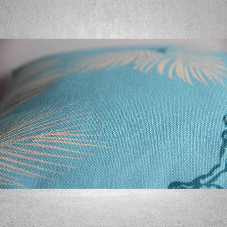 May include: Close-up of a square, light blue pillow with a beige palm frond pattern. The pillow has a textured fabric and a tropical theme. The design is visible on the front of the pillow.