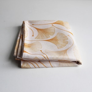 Może przedstawiać: Złożona biała tkanina z powtarzającym się wzorem złotych liści miłorzębu. Liście mają szczegółowy, żyłkowany wygląd i są ułożone na neutralnym tle. Tkanina jest starannie złożona, prezentując wzór.