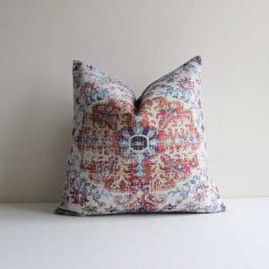 Könnte beinhalten: Dekoratives Zierkissen mit einem verzierten Muster in Orange, Lila, Blau und Weiß. Das Kissen hat ein zentrales Medaillon-Design und einen Vintage-Look, passend für die Inneneinrichtung.