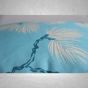 May include: Close-up of a light blue fabric with a repeating pattern of white palm fronds and a dark blue tree branch. The fabric has a textured appearance, suggesting a woven material. The design evokes a tropical theme.