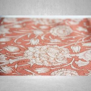May include: Close-up of a coral-colored fabric with a floral pattern. The design features white stylized flowers and leaves. The fabric has a textured appearance, suggesting a woven material.