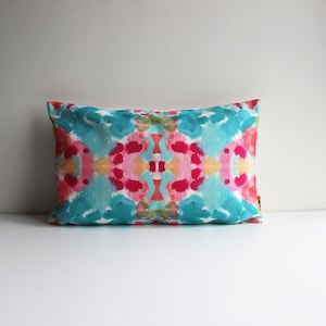Pode incluir: Uma almofada retangular com um design abstrato colorido. A almofada apresenta um padrão simétrico de formas de aquarela rosa, azul e amarela.