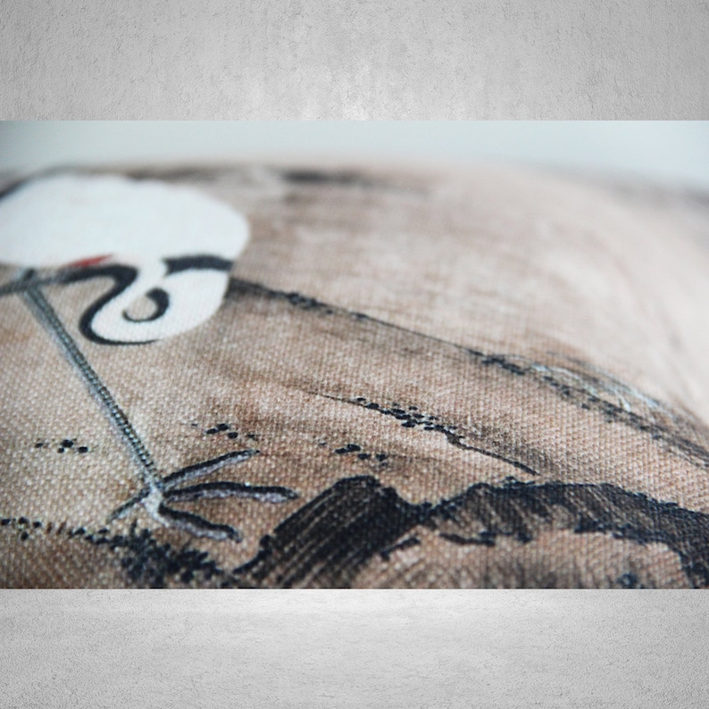May include: A close-up of a beige pillow with a black and white crane design. The crane is depicted in a traditional Japanese style, with long, elegant legs and a long, curved beak. The pillow is made of a soft, textured fabric.