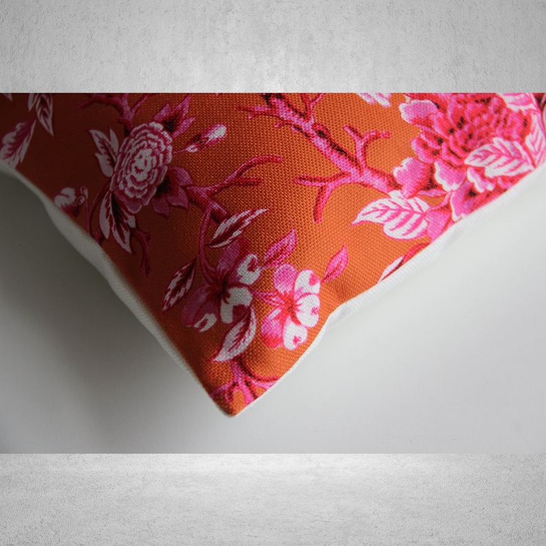 May include: A close-up of a pillow with a floral pattern. The pillow is orange with pink flowers and green leaves. The pillow has a white border.