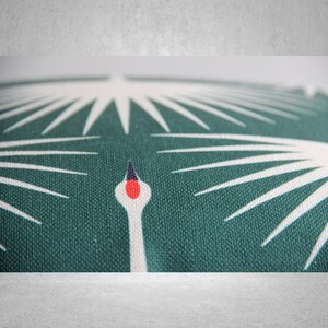 May include: Close-up of a green fabric cushion with a white stylized crane design. The crane's head is red and black.