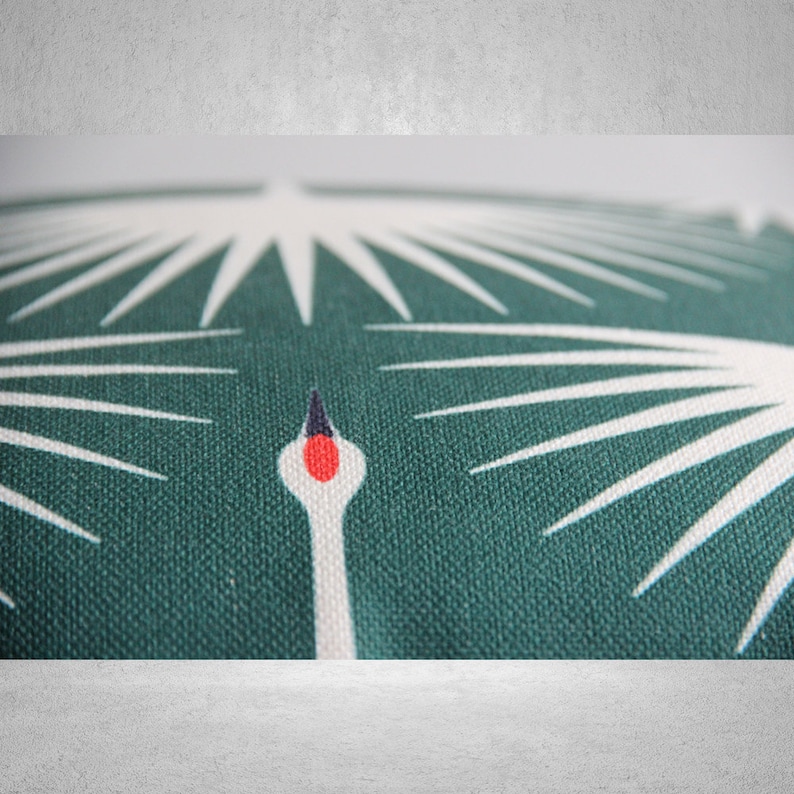 May include: Close-up of a green fabric cushion with a white stylized crane design. The crane's head is red and black.