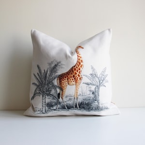 Könnte beinhalten: Ein weißes Kissen mit einer Schwarzweißillustration einer Giraffe, die in einer Dschungelkulisse steht. Die Giraffe ist braun und hat einen langen Hals. Die Dschungelkulisse umfasst Palmen und andere Pflanzen.
