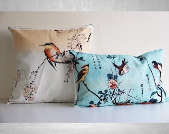 May include: Two decorative throw pillows with a Japanese-inspired design featuring birds and flowers. The larger pillow has a cream background with a light orange bird and pink flowers. The smaller pillow has a light blue background with a yellow bird, a pink flower, and a dark blue bird.