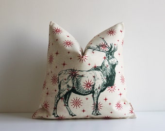 May include: A decorative pillow with a cream-colored background featuring a green illustration of a deer. The pillow is adorned with a pattern of red starbursts. The pillow is a square shape and is placed on a white surface.