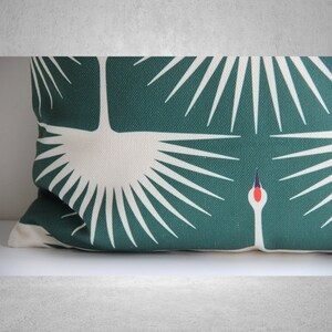 May include: A green and white throw pillow with a stylized design of a crane with its wings spread. The crane is white with a red beak and black eyes. The background is a deep green.