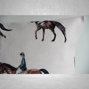 May include: A white cushion with a brown horse and rider pattern. The cushion is made of a textured fabric and has a subtle sheen.