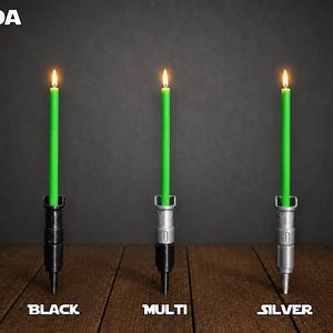 May include: Three lit green taper candles held in vertical holders shaped like lightsaber hilts. The left holder is black, the center is a multi-colored black and silver, and the right is silver. The text YODA appears at the top left, with BLACK, MULTI, and SILVER labels below each candle.