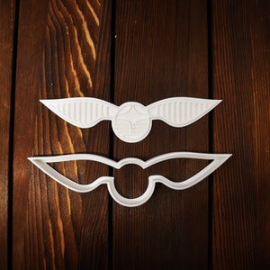 Könnte beinhalten: Zwei weiße Ausstecher in Form des Goldenen Schnatz, ein fiktives Objekt aus der Harry-Potter-Reihe. Einer ist massiv, der andere ein Umriss. Sie liegen auf einer dunklen Holzoberfläche.