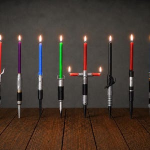May include: Eight lit taper candles in various colors including red, purple, blue, green, and black, each held in a metallic, cylindrical base designed to resemble a sci-fi weapon handle. One candle features a cross-guard design with two side flames.